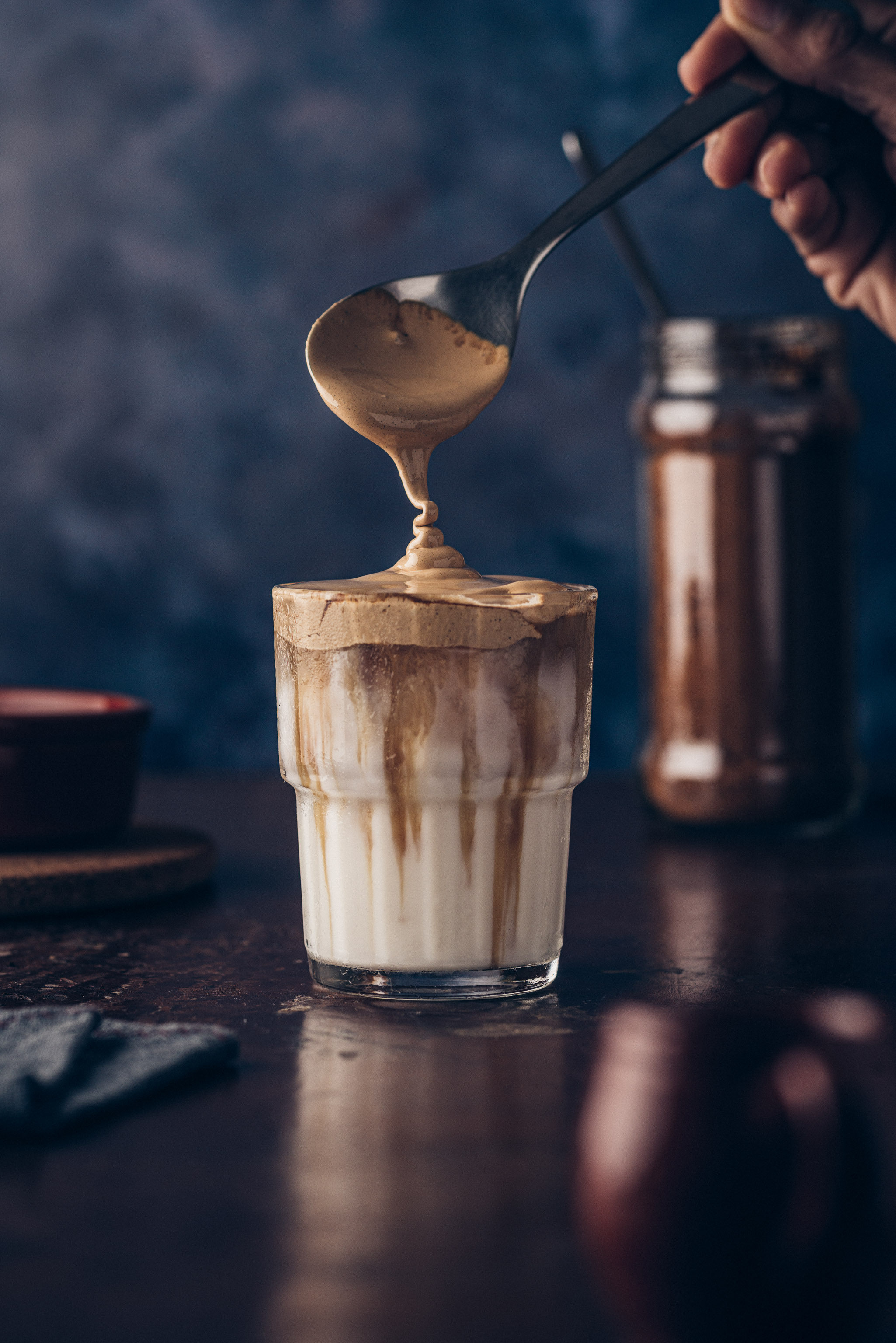 A spoon scooping whipped dalgona coffee onto a glass of iced milk, with a jar of ingredients in the background.